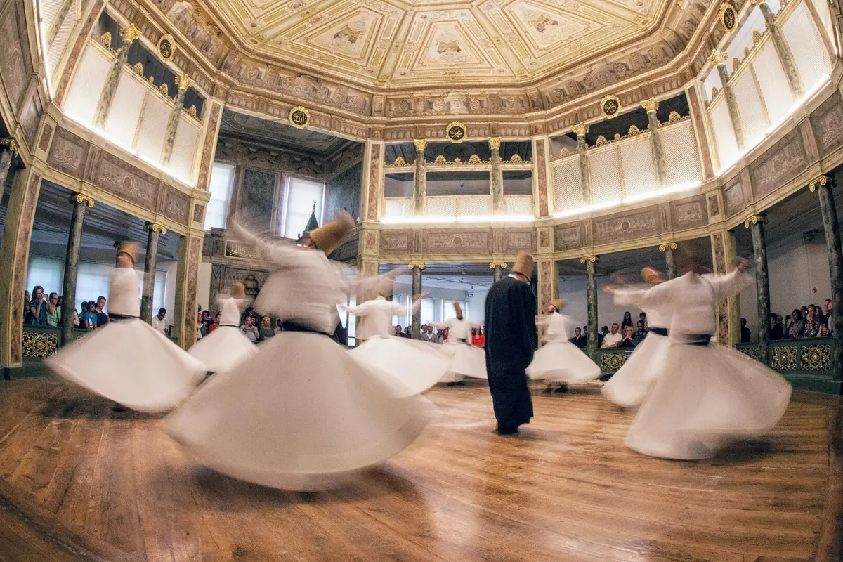 Semazens in white skirts and tall hats whirling on a wooden floor in a round hall with a gilded octagonal ceiling and spectators along the edge.