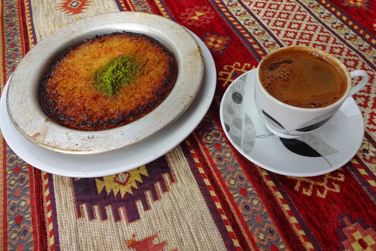 A round golden künefe topped with pistachio beside a cup of Turkish coffee on a red patterned cloth.