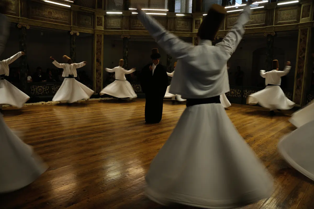 A semazen with arms open turning in the foreground of a dim wood-floored hall, with other dancers beyond.