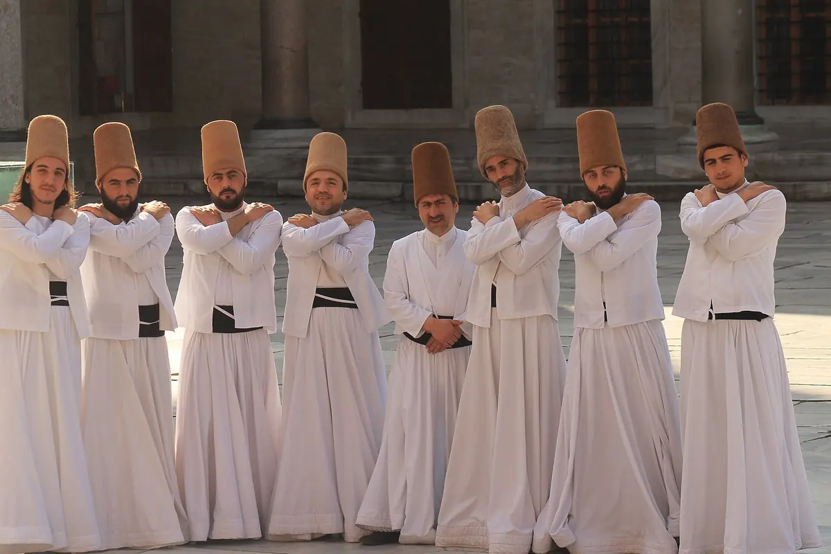 Eight semazens in white robes and tall brown felt hats standing in a row with their arms crossed over their chests.