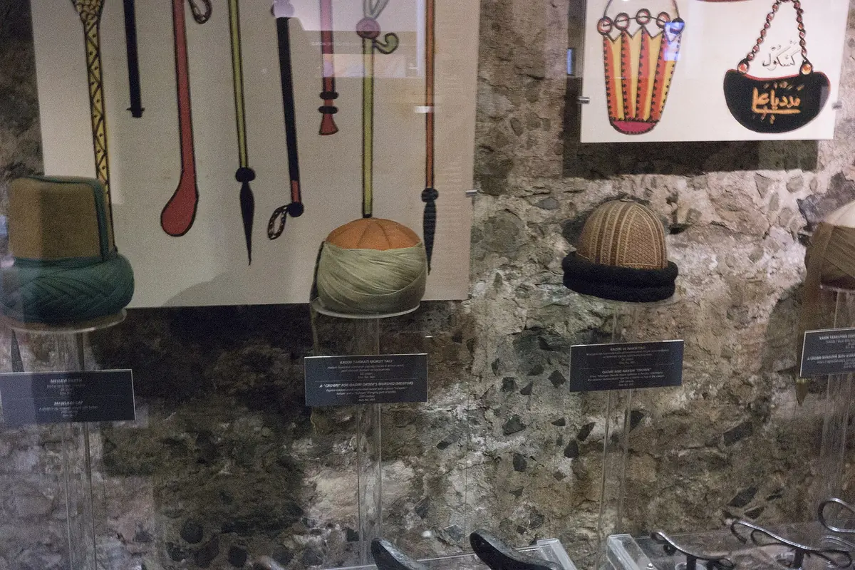 Felt sikke hats in several colours displayed on a stone wall beside printed panels in a museum.